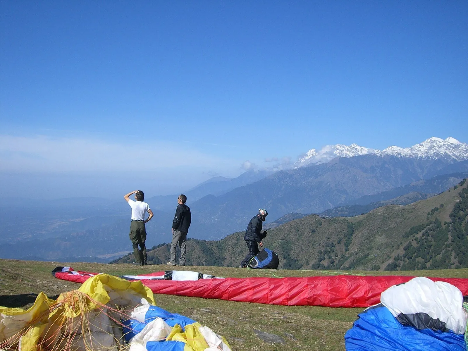 Pilots at the Billing takeoff site at 2,400 m, gliders laid out on grass with Dhauladhar snow peaks across the valley